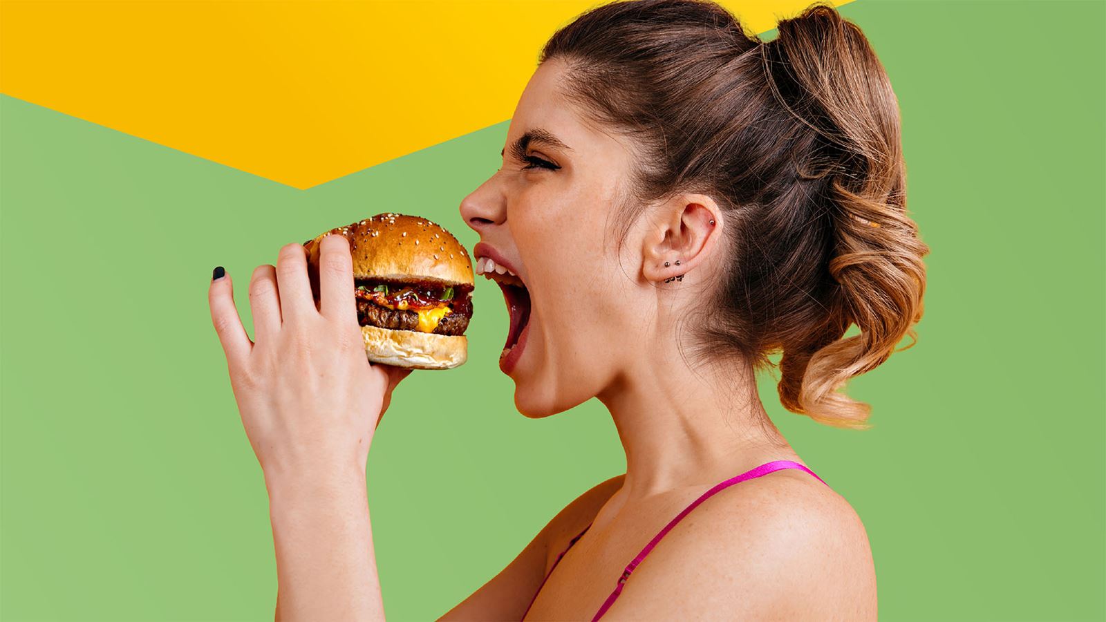 Junge Frau beisst mit Vorfreude in einen saftigen Burger (mit gelbem Designelement oberhalb)