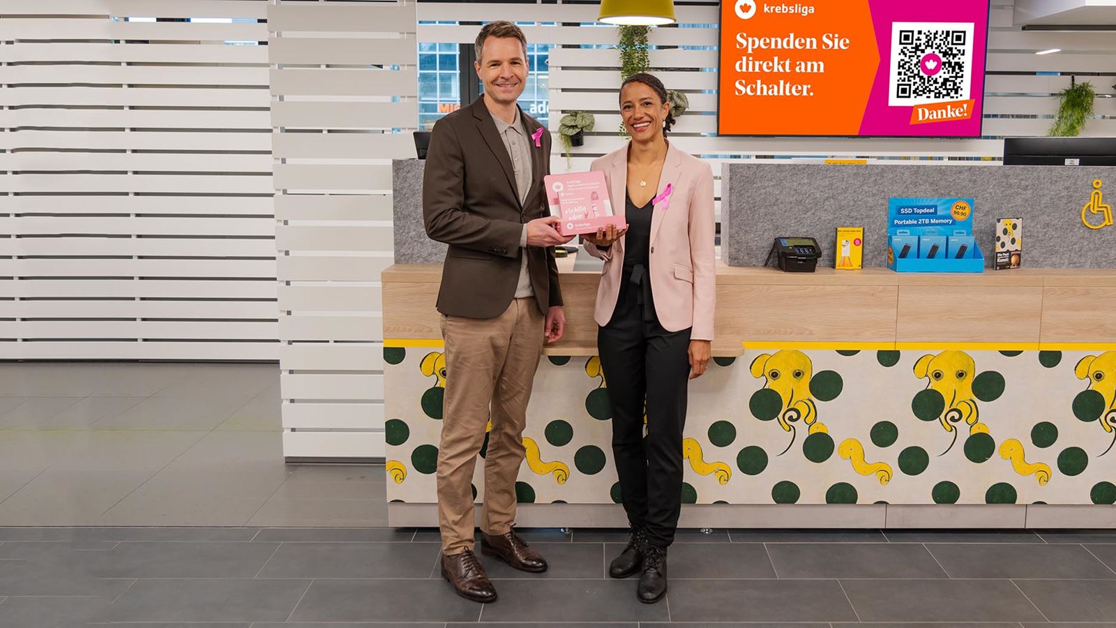 Oliver Egger, responsable Marché des médias et marché publicitaire Swiss Post Advertising, et Mirjam Lämmle, CEO de la Ligue suisse contre le cancer, présentent les rubans roses dans une filiale de la Poste.