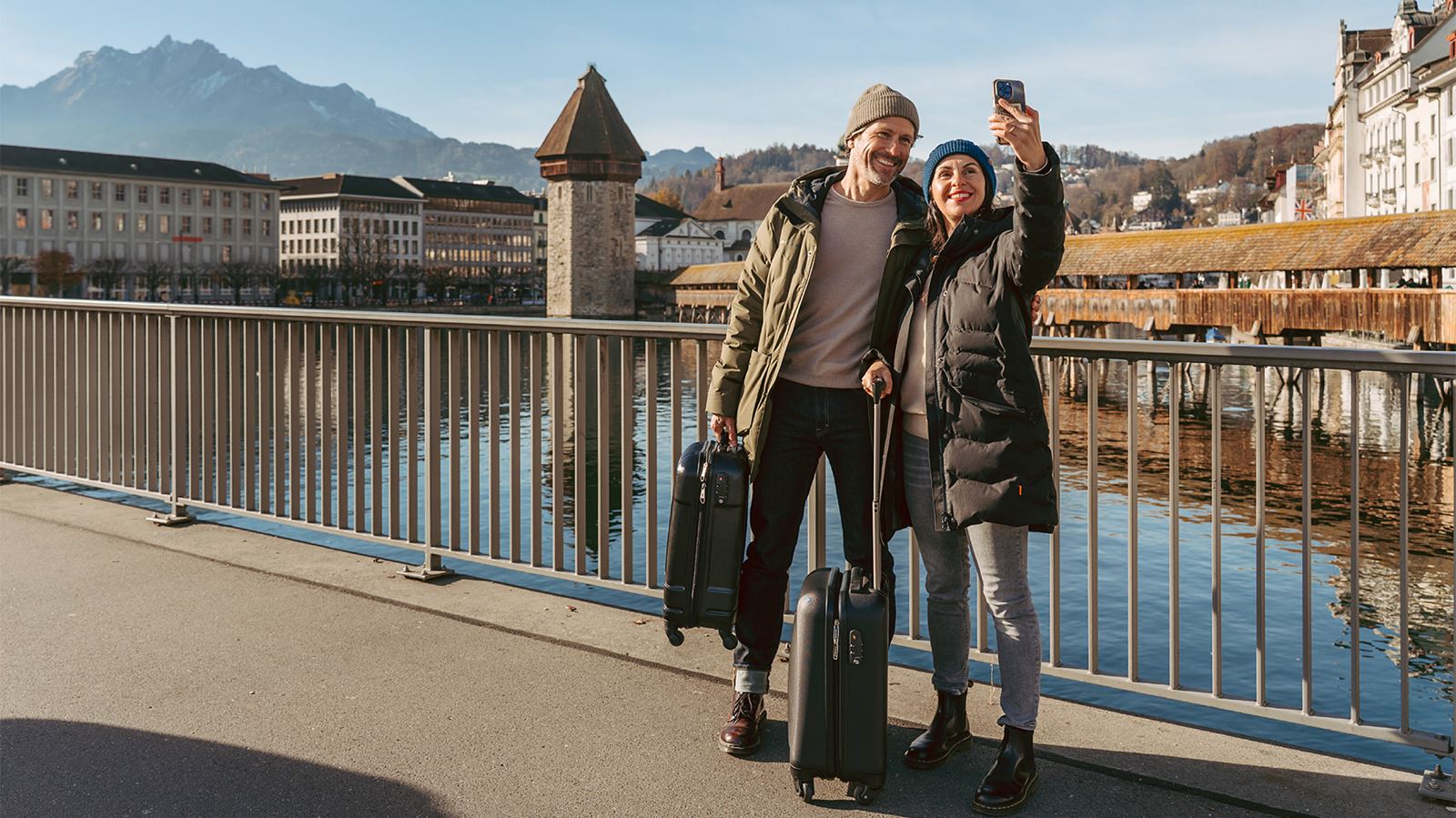 Una coppia di viaggiatori con le valigie si scatta un selfie dal marciapiede di un ponte di Lucerna con il Kapellbrücke sullo sfondo. 