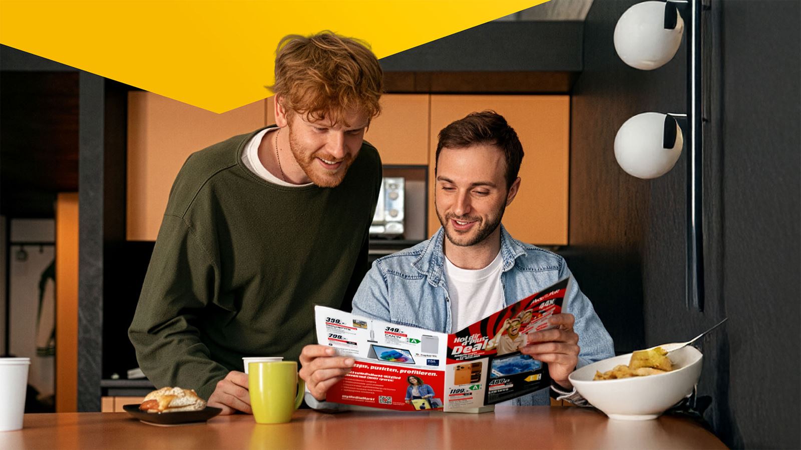 Due giovani uomini studiano un catalogo di prodotti di MediaMarkt a colazione (con elemento di design giallo sopra)
