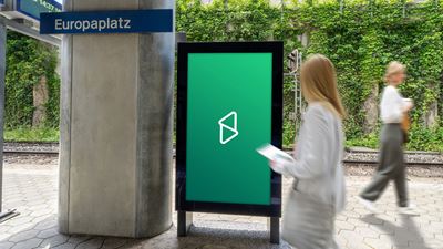 Digitaler Werbescreen an einem Bahnhof mit vorbeigehenden Passantinnen; im Hintergrund sind Gleise und eine begrünte Mauer zu sehen.