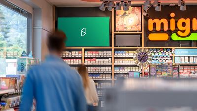 Digitaler Werbescreen im Verkaufsraum eines Kiosks oder Convenience-Shops; im Vordergrund sind unscharfe Kundschaft und Regale mit Produkten zu sehen.